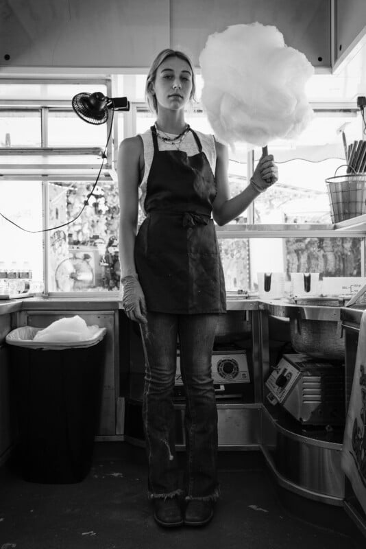 A person stands in a kitchen holding a large cotton candy. They wear an apron over a white shirt and jeans. The kitchen has various equipment and containers, with a trash bin and cotton candy machine visible. Black and white image.