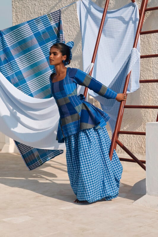 A woman in a blue plaid outfit stands on a stone surface, leaning against a ladder. Plaid and checkered fabric hang in the background, creating a dynamic scene. The setting appears to be outdoors on a sunny day.