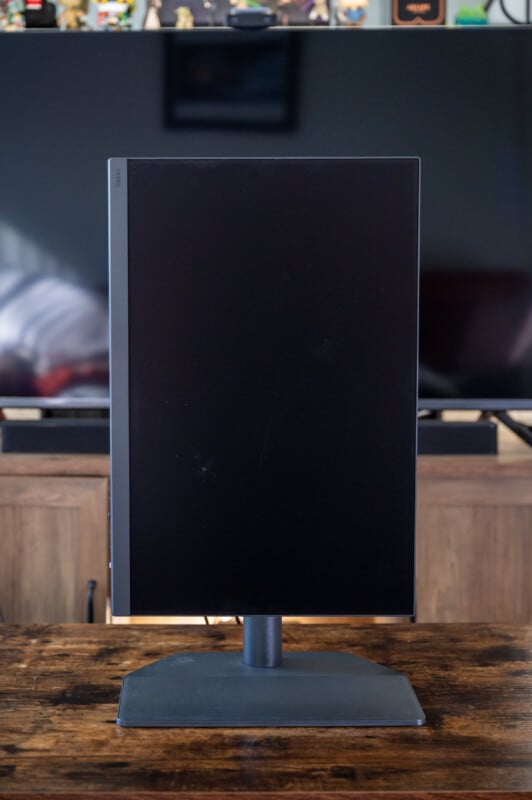 A black computer monitor is rotated to a vertical position on a wooden desk. Behind it is a modern entertainment setup with a large flat-screen TV and shelves with various decorations.