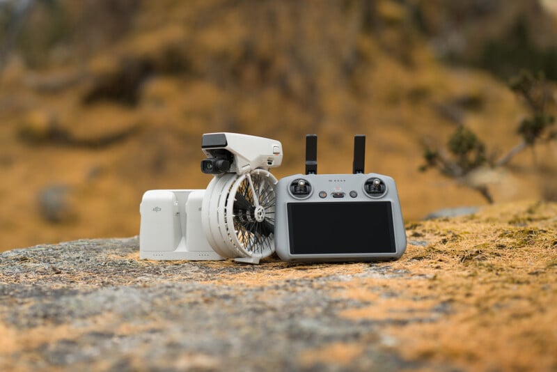 A white drone and its gray remote controller rest on a rocky surface in an outdoor setting with a blurred background of golden grasses and shrubs. The controller has a screen, and the drone features a gimbal camera.