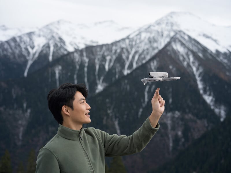 A person in a green jacket smiles while balancing a drone on their finger. Snow-capped mountains are visible in the background under a cloudy sky.