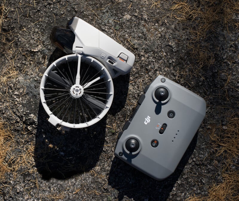 A white drone with a circular propeller guard and a gray remote control rest on dry, textured ground scattered with pine needles.