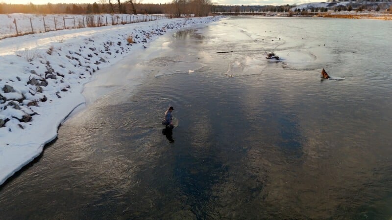 A person stands in a partially frozen river with snowy banks under a cloudy sky. The landscape is lined with trees and a distant hill. Ducks float nearby, adding to the winter scene.