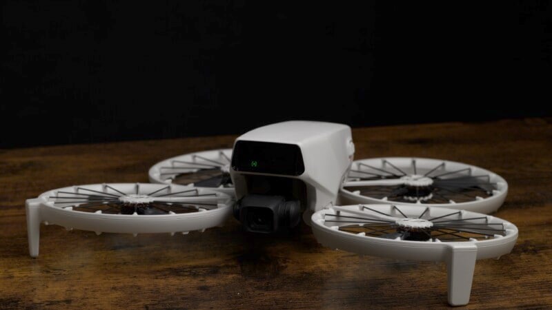 A white quadcopter drone with propeller guards sits on a wooden surface against a dark background. Its compact design and camera are visible, positioned for takeoff or landing.