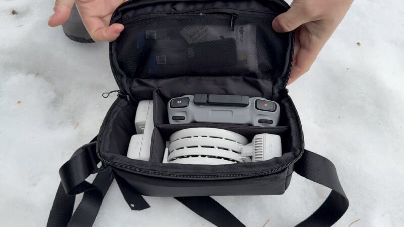 A person holds an open black carrying case on snow, revealing a compact, folded drone inside. The case has mesh pockets and a strap, with hands adjusting the contents.