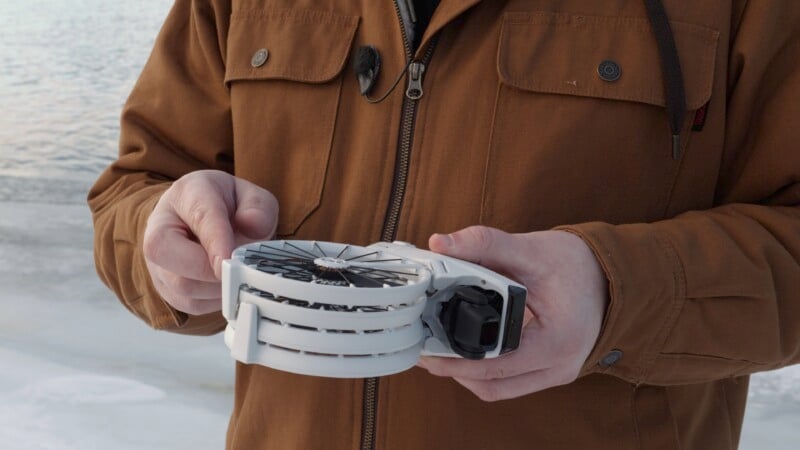 A person in a brown jacket holds a foldable drone near a snowy, icy landscape by the water. The drone is compact and white, with visible propellers.