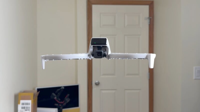 A white drone hovers indoors in front of a closed door. The drone's camera is visible, and there are some objects and a partial view of a desk in the background.