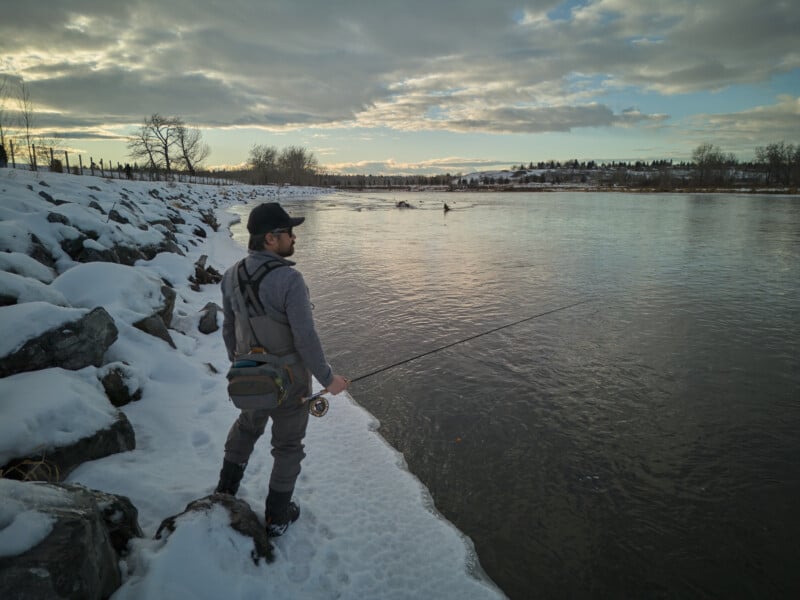 A person in winter clothing is fishing by a snow-covered riverbank under a cloudy sky. The river flows gently, surrounded by leafless trees and distant hills. The peaceful scene captures the essence of winter fishing.