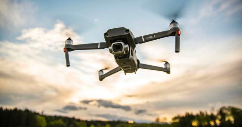 A drone flies in the air against a backdrop of a scenic sunset. The sky features vibrant hues of orange and yellow, with a few clouds. The landscape below shows a forested area and a distant horizon.