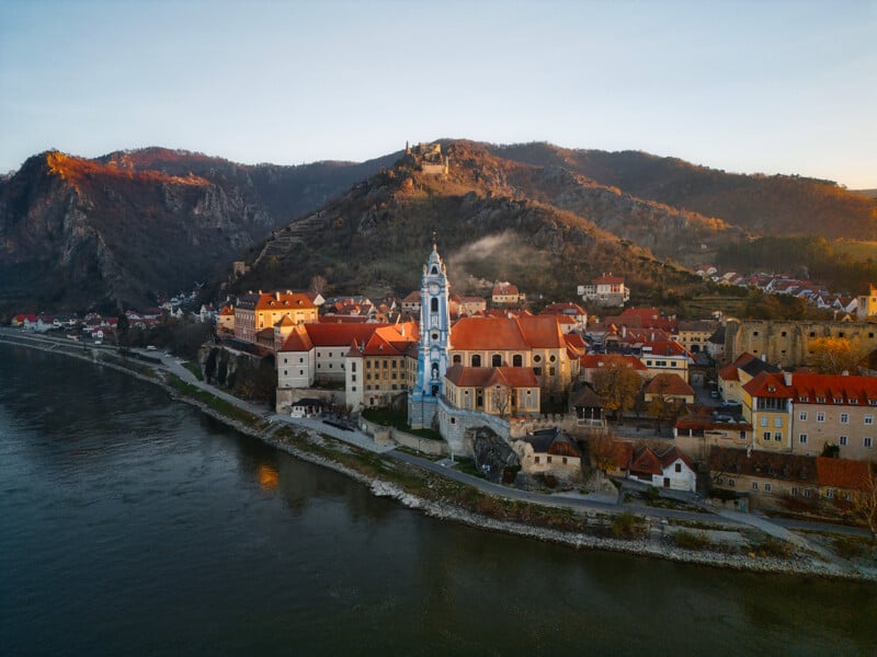 A picturesque view of a historic town with a prominent blue church tower, surrounded by traditional buildings and nestled along a river. The backdrop features hills with a castle ruin under a clear, warm sky.