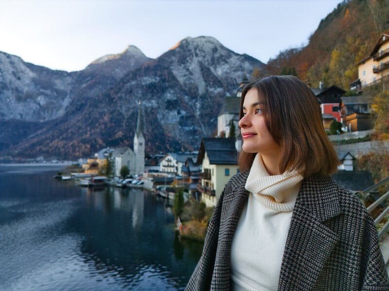 A woman with shoulder-length brown hair and a checkered coat smiles while standing near a scenic lakeside village, surrounded by mountains. The view includes quaint buildings and a serene lake, with an overcast sky in the background.