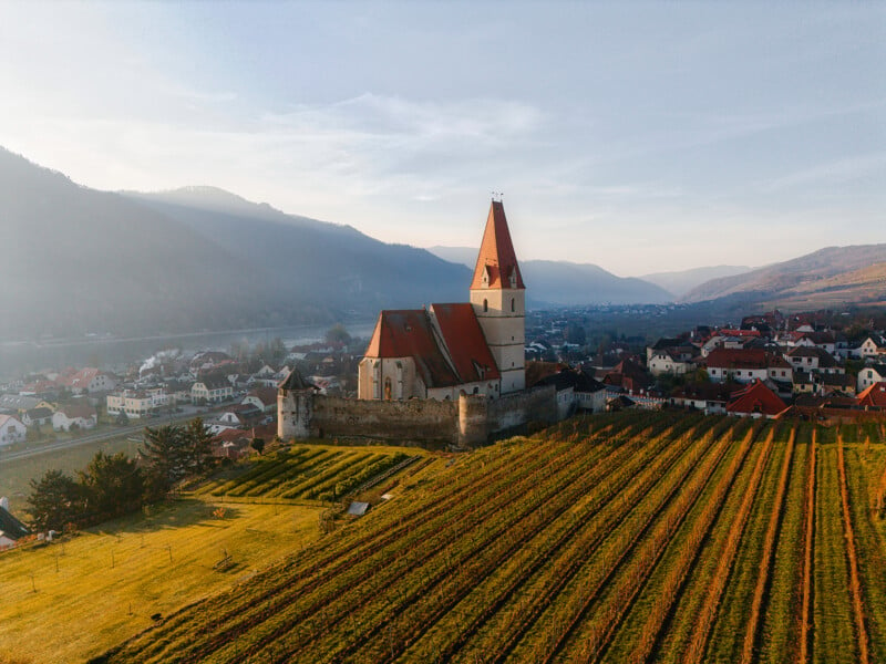 Aerial view of a picturesque village with a central church featuring a tall red-roofed tower. Surrounding it are lush vineyards, quaint houses, and rolling hills under a clear blue sky.