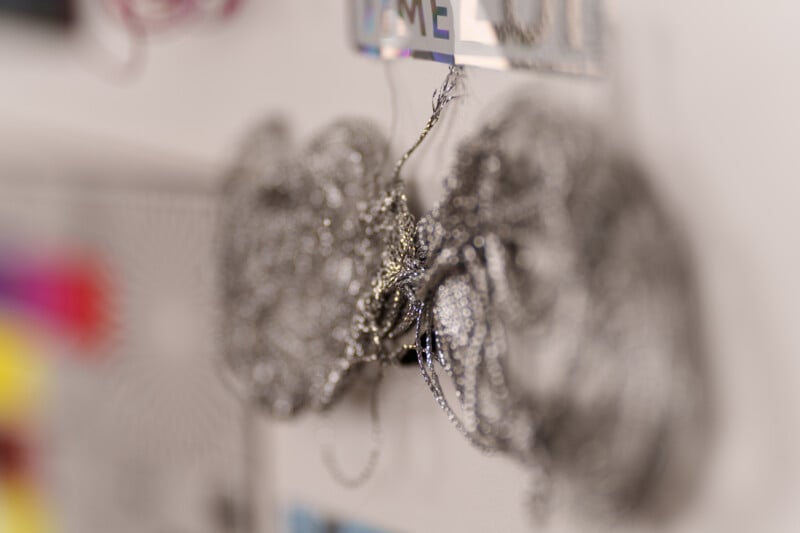 Close-up of tangled metallic chains or wires hanging on a wall. The background is blurred with soft colors and patterns, hinting at other objects or decorations nearby.