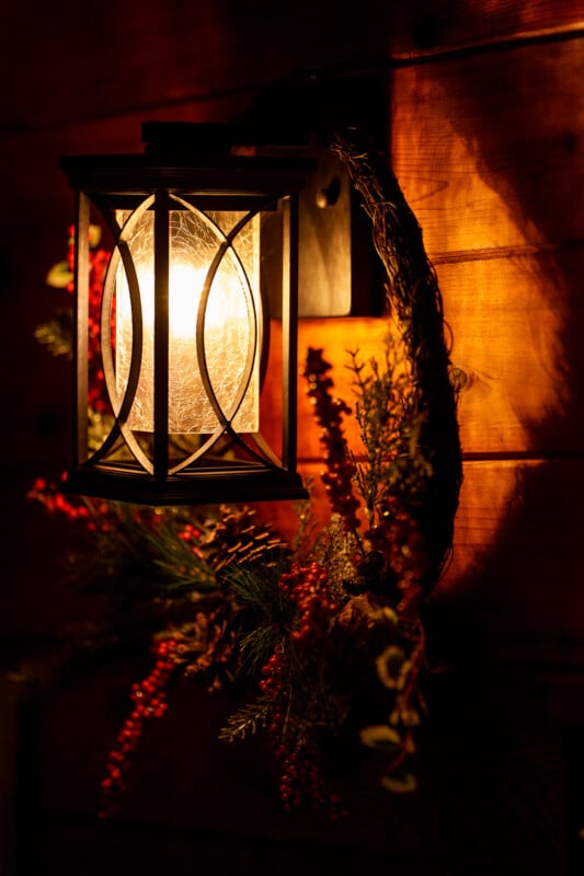 A softly glowing wall lantern with a decorative glass enclosure, mounted on a wooden wall. Surrounding the lantern is a festive wreath adorned with red berries and greenery, casting warm light and creating a cozy ambiance.