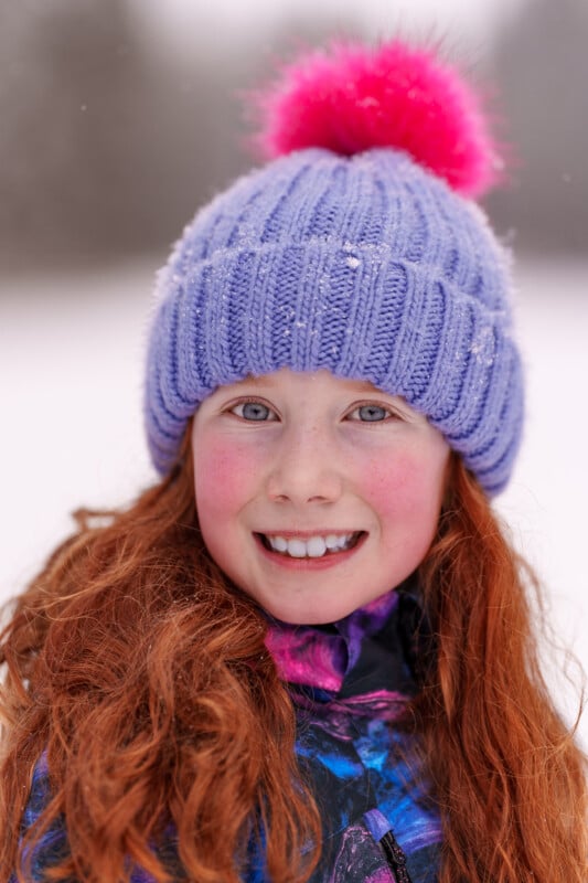 Young girl with red hair smiling, wearing a purple knit hat with a pink pom-pom and a colorful winter jacket, standing in a snowy setting. Snowflakes dot her hat, and her cheeks are rosy.