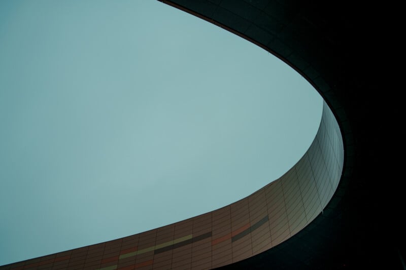 An architectural photograph showcasing a curve of a modern building against a clear sky. The structure features a smooth, sweeping design with a mix of dark and earthy tones.