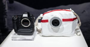 A Nikon camera sits next to a white spherical camera with red accents, resembling a design inspired by space gear, on a white surface. The background is dark with a moonlike texture.