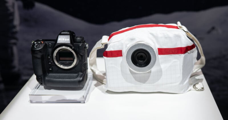 A Nikon camera sits next to a white spherical camera with red accents, resembling a design inspired by space gear, on a white surface. The background is dark with a moonlike texture.