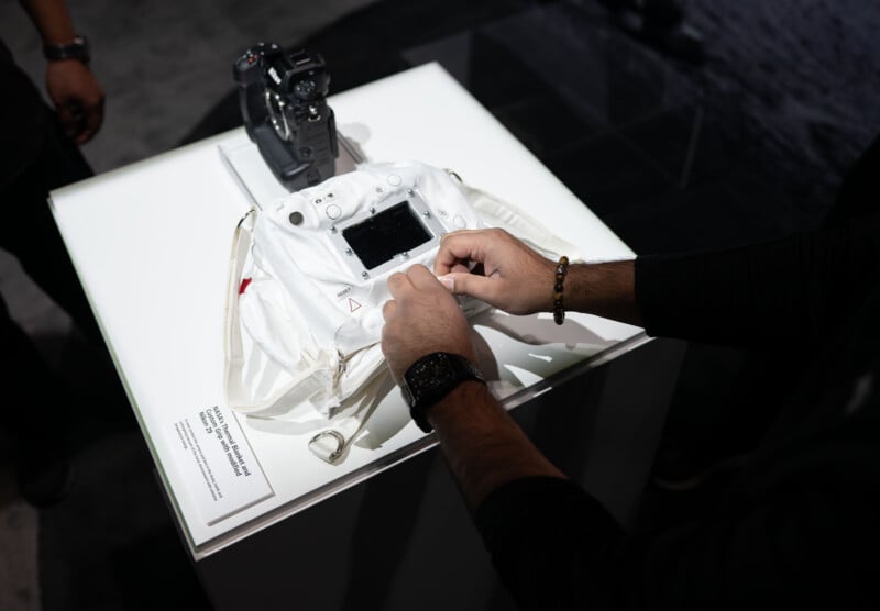 A person handles astronaut gloves on a display table beside a camera. The gloves have a control panel, and there is an informational placard on the table. The background is dimly lit.