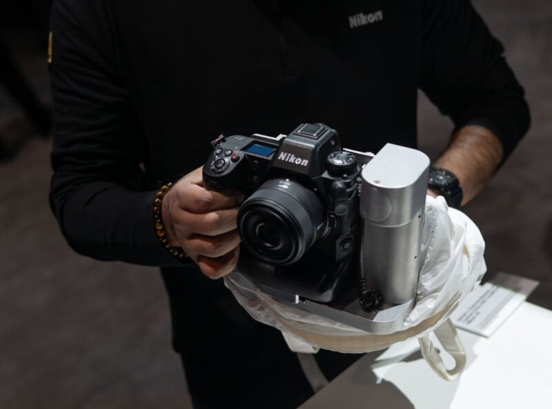 A person holds a Nikon camera mounted on a stabilizing device, which is resting on a white cushion. The person is wearing a long-sleeve black top. The setup is showcased on a table in a dimly lit environment.