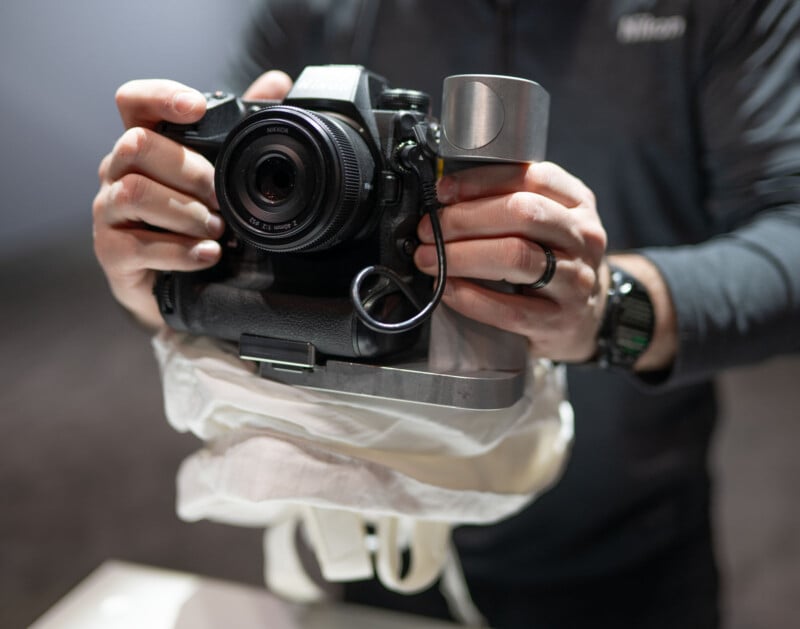 A person holding a camera modified with a custom metal grip and a cable attached, wearing a black long-sleeve shirt and a wristwatch. The camera is partially supported by a white fabric sling or harness.