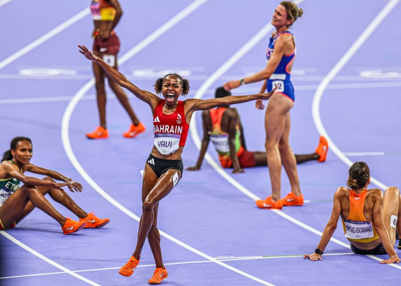 A track athlete from Bahrain exults with arms wide open on a violet track surface. Other competitors are seated or sprawled on the ground, exhibiting weariness, dressed in colorful clothing and footwear.