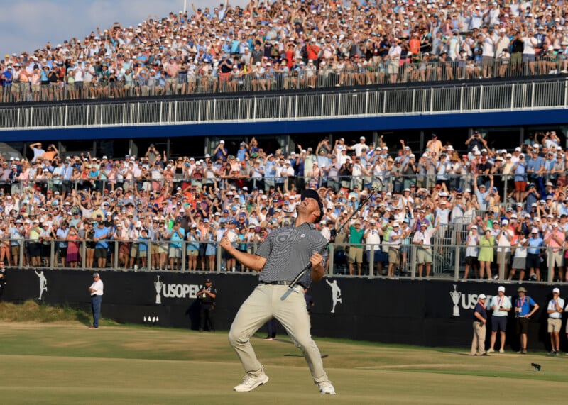 A golfer exclaims in joy on a green during a competition. He holds a putter and appears overjoyed. Behind him, a large audience of spectators fills the stands, cheering and capturing images, with a "US Open" sign evident.