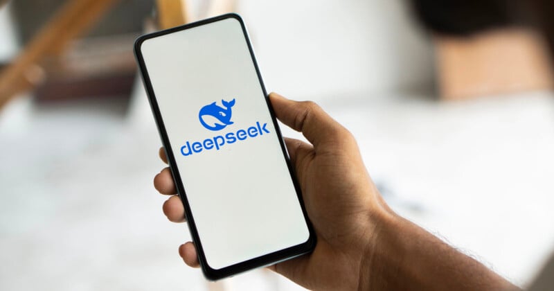 A person holds a smartphone displaying the "deepseek" logo, featuring a stylized rhinoceros head. The screen background is white, and the setting appears to be indoors with a blurred background.