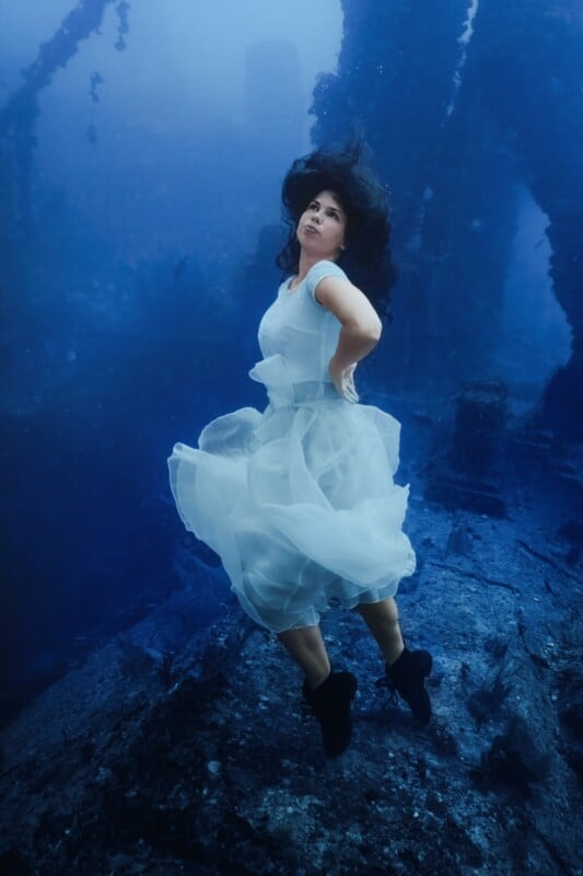 A person wearing a flowing white dress and black shoes is underwater, surrounded by a blue ocean and the structures of a sunken ship. Their hair floats around them as they gaze upwards.