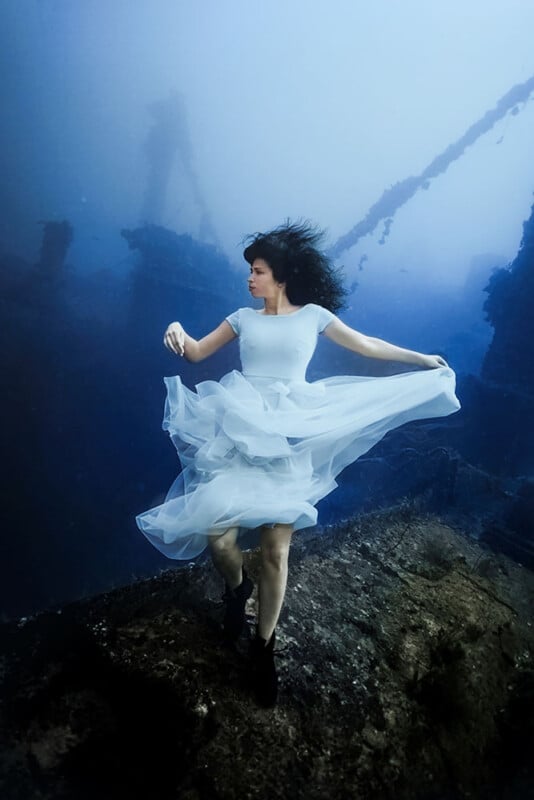 A woman in a flowing white dress is underwater, surrounded by the remnants of a shipwreck. Her dark hair floats around her as she stands on a piece of wreckage, creating an ethereal and dreamlike scene.