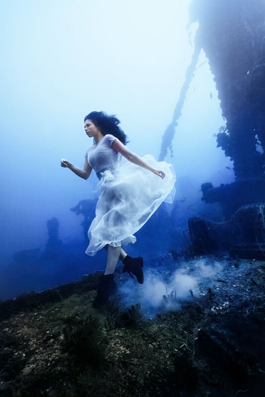 A woman in a flowing white dress walks underwater amid a sunken shipwreck. Her dark hair moves freely, and her dress billows around her. The background shows a silhouette of the ship surrounded by blue water.