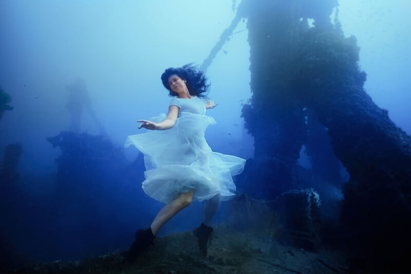 A woman in a flowing white dress dances gracefully underwater, surrounded by dark, shadowy structures. Her hair billows around her as she moves through the blue-tinged water, creating an ethereal and dreamlike atmosphere.