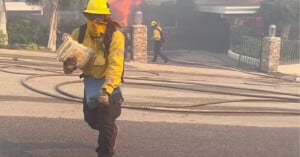 A firefighter in yellow protective gear and helmet carries a container while walking on a smoky street. In the background, another firefighter stands near a building, with flames visible behind them and hoses stretched across the ground.