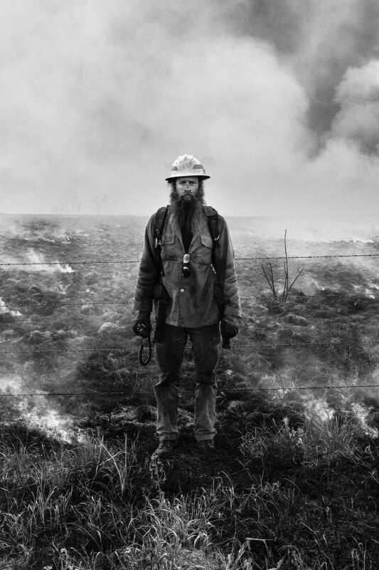 A firefighter wearing protective gear, including a helmet and gloves, stands amidst smoke and charred ground. He is looking directly at the camera, with a background of thick smoke and sparse vegetation. The image is in black and white.