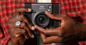 A person holding an Instax Wide Evo instant camera with both hands. The person is wearing a red checkered jacket and silver rings. The camera lens is centered, and the Instax logo is visible at the top.