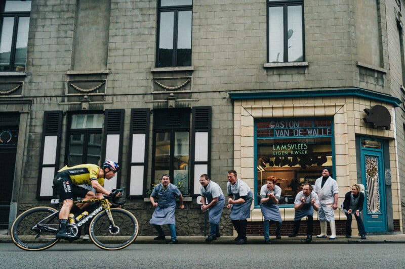 A cyclist in a yellow and black uniform rides past a group of cheering people in aprons outside a building. Some appear to be clapping, while others are leaning forward, watching with excitement.