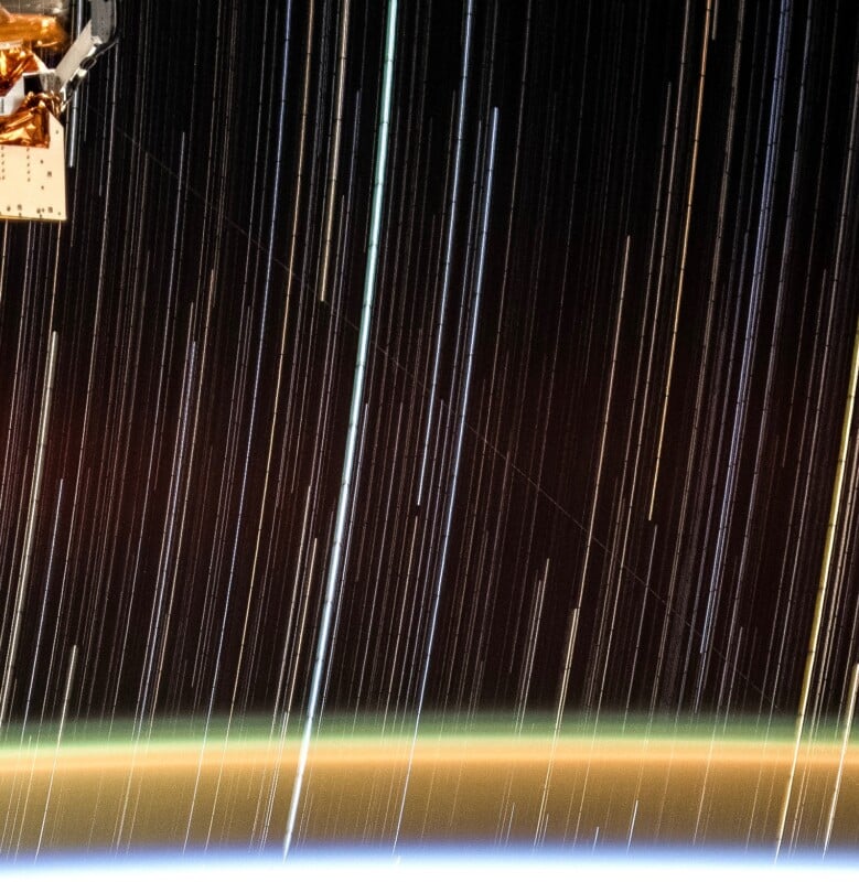 A view from space showing star trails above the Earth's horizon, with part of a spacecraft or satellite equipment visible in the upper left corner. The horizon glows with a gradient of colors from blue to orange.