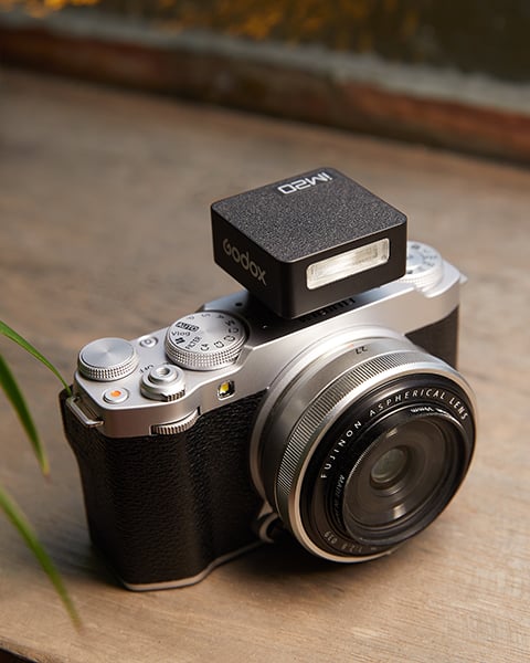 A vintage-style black and silver camera with a Godox flash mounted on top, resting on a wooden surface. The camera has a large lens and traditional control dials, with a small plant visible to the side.