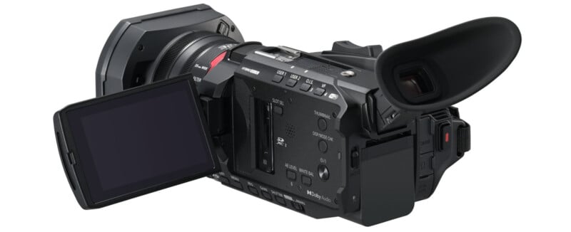 A black professional video camera with a flip-out LCD screen, various controls and buttons, and a large lens. The camera is angled to show the viewfinder, lens, and screen.