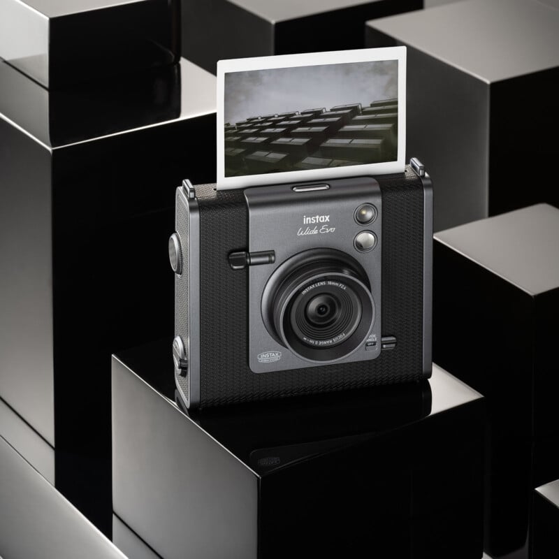 A black instant camera sits on a glossy, black geometric surface. It has a photograph emerging from the top, partially displaying an image. The camera features a textured grip and a lens at the front.