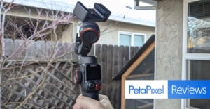 A person holds a Hohem camera gimbal outdoors near a wooden fence. The gimbal features a screen and buttons and is positioned against a backdrop of a house and leafless branches. The image includes a "PetaPixel Reviews" logo.