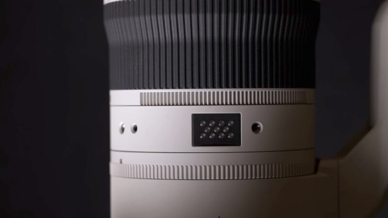 Close-up of a camera lens with a ribbed focus ring and small black section featuring electronic contacts. The lens body is in a sleek, light color against a dark background, showcasing its detailed texture and design.