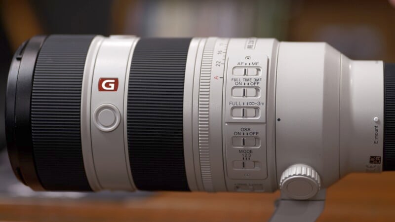 Side view of a white telephoto camera lens with a black rubber focus ring and various control switches. The "G" logo is visible, indicating it's a high-performance lens. The background is blurred.