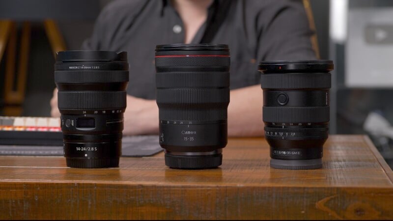 Three camera lenses of varying sizes are displayed on a wooden table. The person in the background is blurred, emphasizing the lenses. The lenses include a Nikkor Z, a Canon RF, and a Sony G.