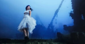 A woman in a flowing white dress stands underwater, surrounded by the remnants of a shipwreck. The water is a striking blue, and her black boots contrast with the ethereal scene. Her hair floats gently, adding to the dreamlike atmosphere.