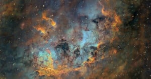 A vivid image of a nebula in space, with swirling clouds of gas and dust in shades of orange, blue, and green. Dotted with bright white and yellow stars scattered throughout the dark, infinite background.