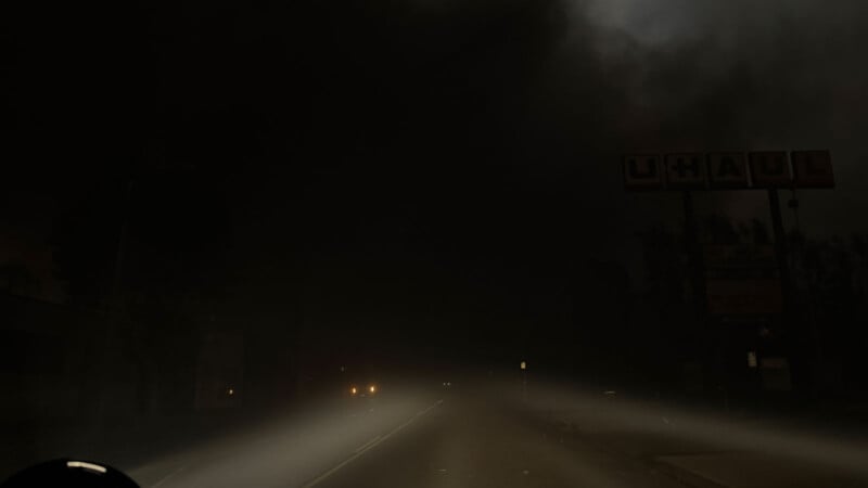 A dark, foggy road at night with dim car headlights visible. A billboard with partially visible letters "UHAUL" stands in the background, surrounded by silhouettes of trees and buildings.