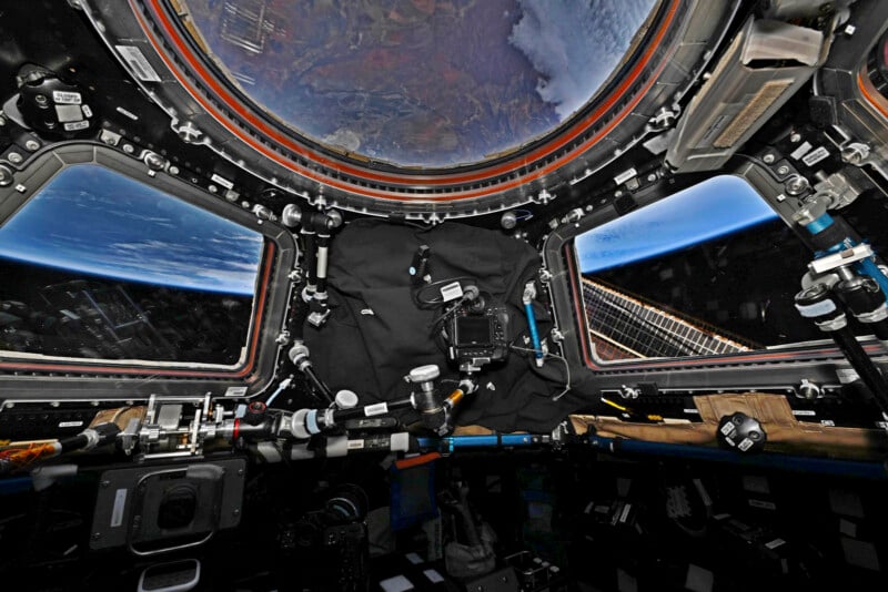 View from the International Space Station's cupola module, showing Earth through large windows. The interior is equipped with various controls, instruments, and cameras. The planet's curve and atmosphere are visible against the blackness of space.