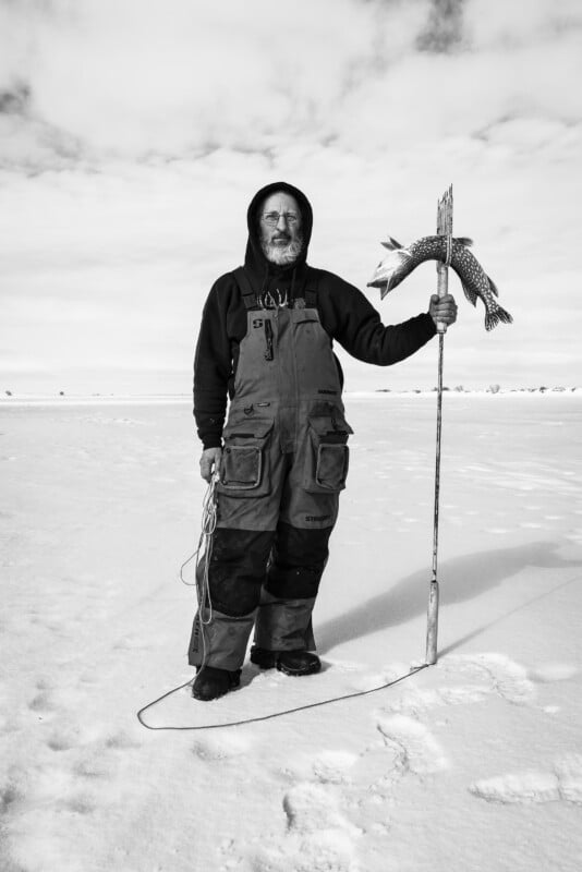 A figure stands in a snowy landscape, wearing overalls and a hoodie. They are holding a staff with a fish sculpture atop it. The sky is overcast, and snow blankets the ground. The scene appears bleak and wintery.