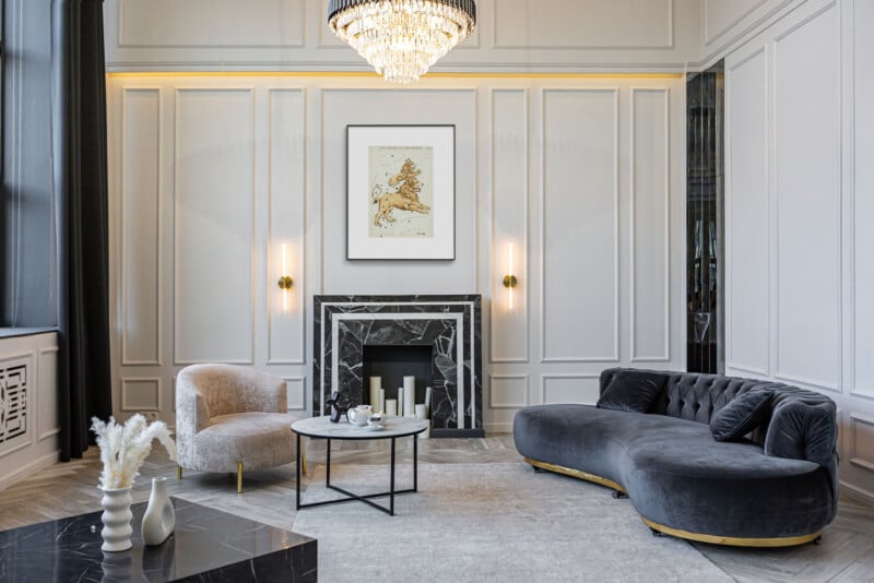 A sophisticated living room with a modern, curved gray sofa, a plush armchair, and a marble coffee table. A decorative fireplace is framed by paneling and wall sconces. A large chandelier hangs from the ceiling, enhancing the elegant ambiance.